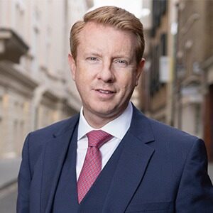 A portrait of Lee Kelly. He is a blond white man wearing a dark suit and vest with a red tie.