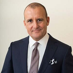 A portrait of Luca Virgilio. He is an older white man wearing a dark suit and gray-red tie.