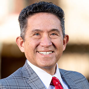 A portrait of Percy Brandon. He is an older man with gelled black hair, a plaid grey suit and a red tie