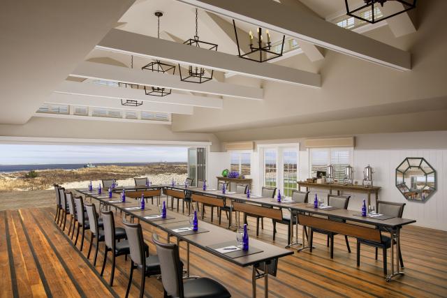 The interior of Chatham Bars Inn. A long table in a horseshoe shape has chairs lined up. The beach is behind, visible through an open wall.