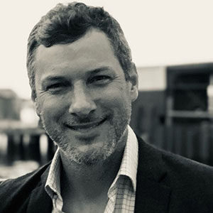 A black and white portrait of Robin Flanz. He is a white man with dark hair, a stubble beard and a dark suit jacket